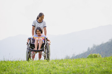 Disabled handicapped child sitting in wheelchair and care helper walking on mountain meadow park in...