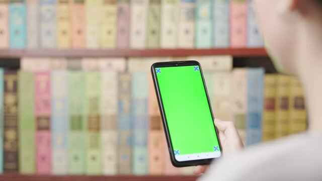 A female's hand holds a smartphone and flips through the pages on a green screen. Close-up. A bookshelf in the background is blurred. Concept of social networks and phone apps.