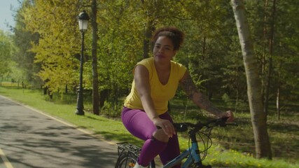 Cheerful positive hipster black female cyclist riding on bike through park, exploring beautiful summer nature, enjoying freedom and active lifestyle while spending leisure during weekend outdoor.