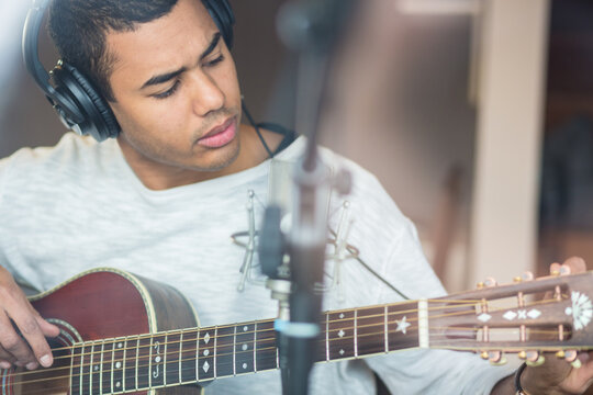 View from outside the window,Afro american young man male musician sitting on chair practicing learning playing guitar using laptop,professional music equipment with microphone and headphone at home