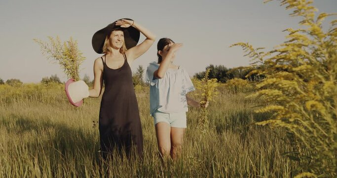 Mother and daughter, walk on a yellow summer lawn under the sun, non urban scene. Girls going straight ahead slowly, looking at horizon. Holding hat in a hand. Nature background, weekend out of city