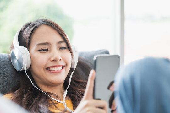 Beautiful Asian Young Woman Wearing Headset And Using Mobile Smartphone. She Leisure On Sofa And Enjoy Watching Device Screen Smart Phone