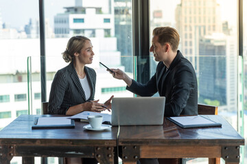 Two business man and woman talkin to each other discussing their business proposal. Both wearing white shirt and black jacket. They are sitting on the wooden table full of glass.