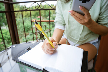 Young caucasian woman writes in a notebook with a pen in casual clothes with a phone in her hand on a summer balcony