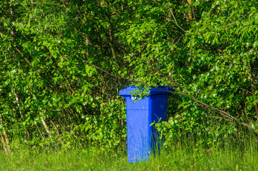 single empty blue waste bin in a public park standing in the bushes