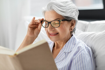 technology, old age and people concept - happy smiling senior woman in glasses reading book in bed...