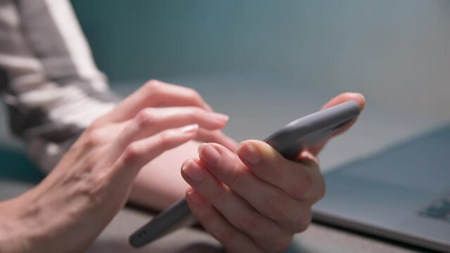 Close-up of a young caucasian woman's hand holding a smartphone. They use the gadget while sitting at the table. Scroll and swipe