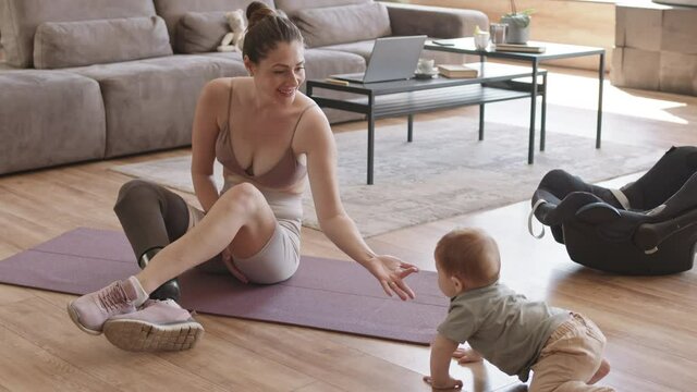 Wide shot of handicapped sporty mommy with leg prosthesis sitting on rubber mat in living room after training and playing with toddler crawling nearby