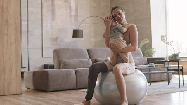 Tilt up of handicapped Caucasian sporty woman with leg prosthesis sitting on fitball at home, holding little son and trying to calm him