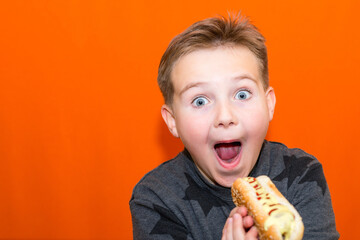 Surprised 10s boy wants to bite a big hot dog.Closeup orange studio background.