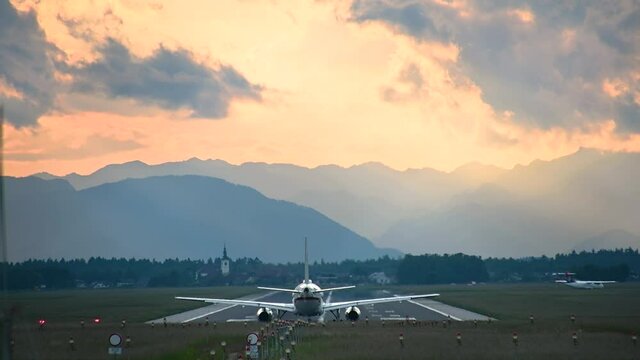 Large passenger airplane standing on the runway against beautiful dramatic sunset at Brnik airport, Slovenia. Aircraft waiting for take-off clearance. View of long runway and Alps mountains