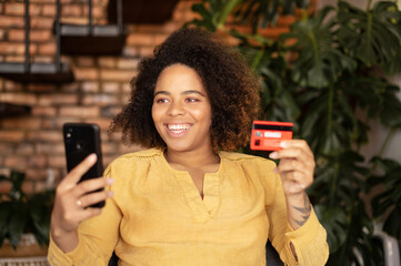 Online payment. Young happy african-American woman holds bank card and smartphone sitting on the armchair at home, making purchase online, ordering food delivery, shopping and paying online