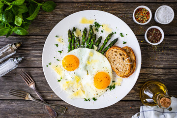 Tasty breakfast. Sunny side up eggs with green asparagus and parmesan served on black plate on wooden table
