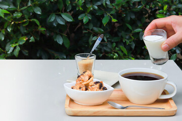 Pour milk into a coffee cup on a white table, where cookies and sugar are placed on a wooden tray at side. Atmosphere in the garden, there is a blurry green leaf in the background.