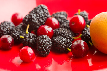 Ripe large black mulberries, peach and red cherries on a red plate, close-up.