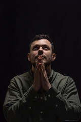 Portrait of a young man clasping his hands in a prayer gesture and looking up against a dark background.