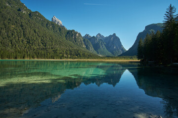 Beautiful mirror reflections in the crystal clear water of Toblacher See lake surrounded by forest and mountains with the Dolomites in the background in Tyrol, Northern Italy. 