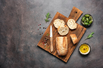 Fresh Italian ciabatta bread on a wooden cutting board with olive oil, olives and herbs, dark rustic background. Top view, flat lay, copy space.