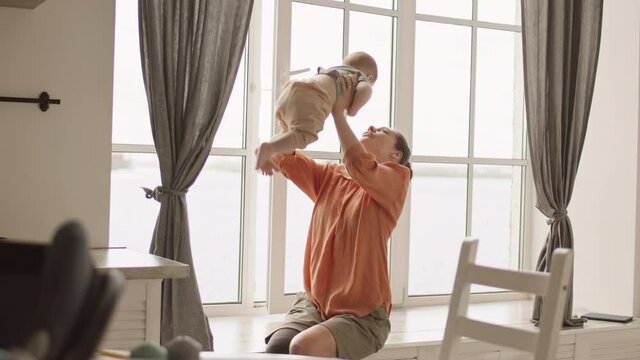 Medium shot of charming Caucasian woman with leg prosthesis sitting on windowsill with cute toddler in her hands, playing with him and then showing view from window