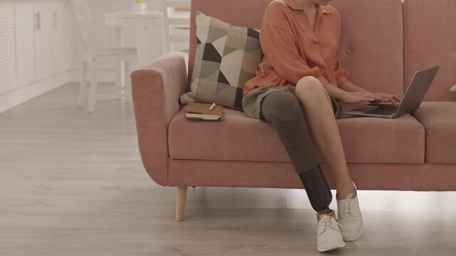 Lockdown of unrecognizable disabled Caucasian woman with leg prosthesis sitting on couch at home, finishing work on laptop, clothing it and going away