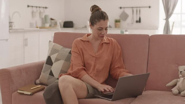 Tracking right of attractive disabled Caucasian woman with leg prosthesis sitting on couch at home and working on laptop
