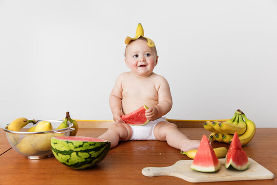 Smiling baby sitting on table wearing banana skin on head holding watermelon piece