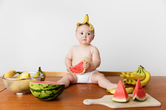Chubby baby sitting on table wearing banana skin on head holding watermelon piece