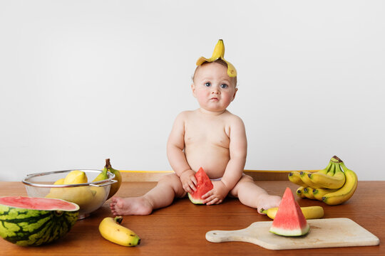 Cute baby sitting on table wearing banana skin on head holding watermelon piece