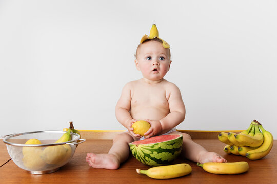 Funny baby sitting on table with fruits wearing banana skin on head