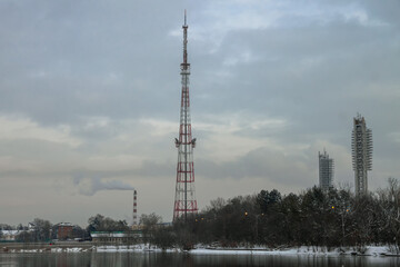 A cell tower, stadium floodlights, and a CHPP chimney against a lake and overcast sky