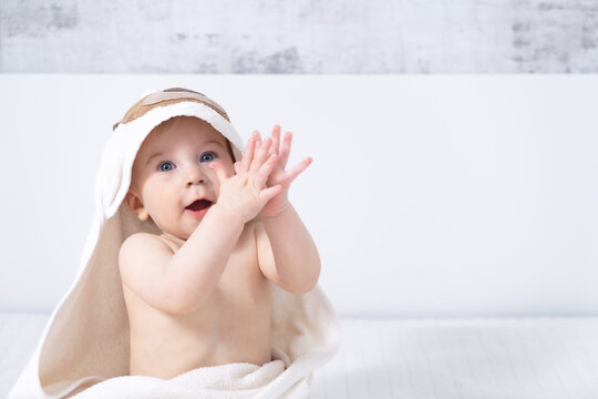Beautiful ten month happy baby girl sitting on bed claping hands indoors in light modern interior with towel after bath. Baby care concept, copy space