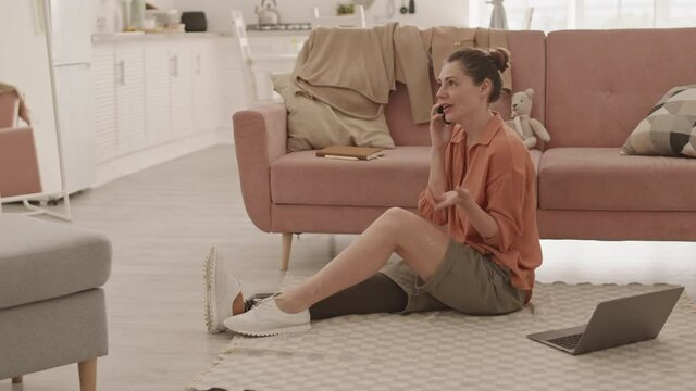 Wide shot of attractive mid adult disabled Caucasian woman with leg prosthesis sitting on floor on carpet in living room and talking on telephone