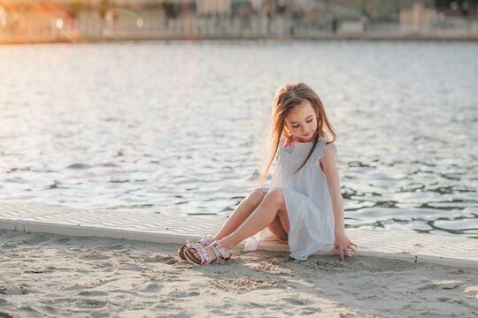 A little cute girl in a white dress sits on the sand on the beach, the child smiles, filmed at sunset, warm colors, summer time, children's recreation and tourism. 