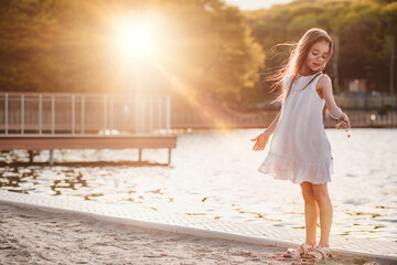 A little cute girl in a white dress sits on the sand on the beach, the child smiles, filmed at sunset, warm colors, summer time, children's recreation and tourism. 