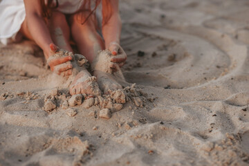 A little cute girl in a white dress sits on the sand on the beach, the child smiles, filmed at sunset, warm colors, summer time, children's recreation and tourism. 