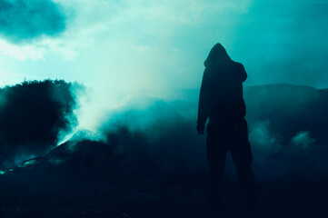 An atmospheric, moody concept. Of a lone hooded figure standing in front a burning pile with smoke covering the area.