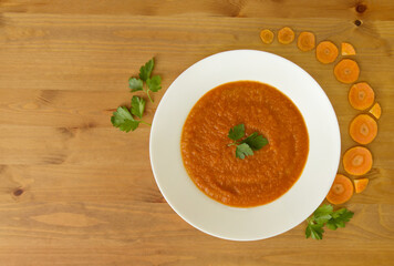 Creamy carrot and pumpkin puree soup on a wooden table decorated with parsley and slices 
of fresh carrots in a white plate on a wooden table top view