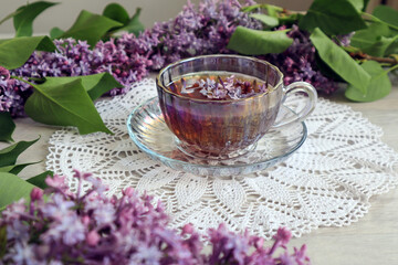 Obraz na płótnie Canvas The concept of a good spring morning. A cup of tea with lilac flowers in it among the lilac branches on a white openwork napkin, side view