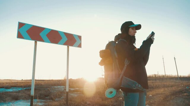 A Young Woman Traveler Uses Her Phone To Choose The Right Road. Hiker Woman With Backpack Choose In A Road Fork Between Two Different Path Directions At The Sunset. Concept Of Choose The Correct Way.