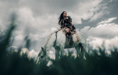 Woman rides on horseback in image of warrior amazon on grass and sky background.