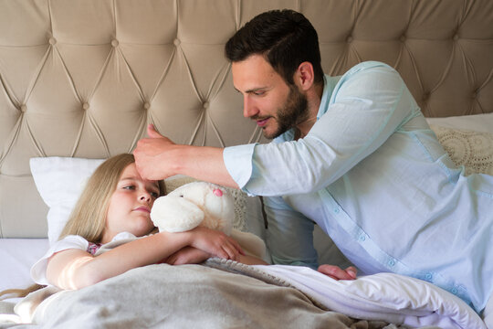 An adorable ill girl is lying in bed with her teddy bear, and the father is checking her temperature. - Powered by Adobe