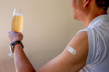 An Asian man is showing plaster on shoulder after coronavirus vaccine.  Man holding a glass of champagne to celebrate being vaccinated.