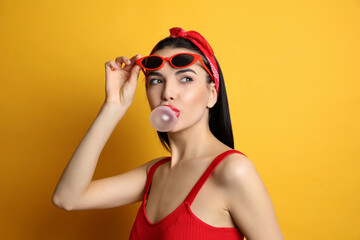 Fashionable young woman in pin up outfit blowing bubblegum on yellow background