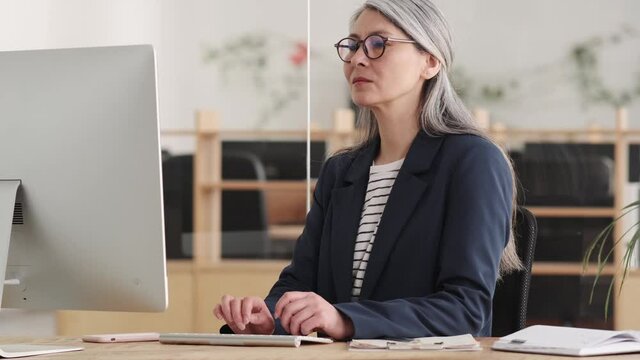 A tired asian woman with gray hair taking off glasses and smoothing the bridge of her nose while sitting at a table in front of a computer in the office