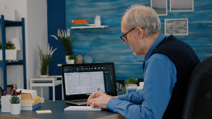 Senior man architect analysing digital prototype with plans from laptop working from home, taking...