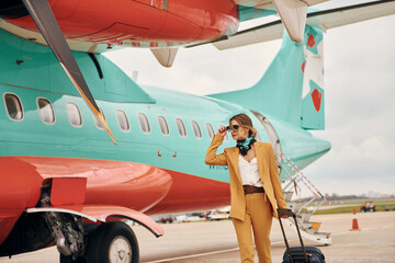 Obraz premium Passenger woman that is in yellow clothes, sunglasses and with luggage is outdoors near plane