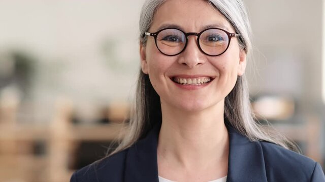 A close-up view of the positive asian woman in a glasses with gray hair doing hello gesture at the camera in the office