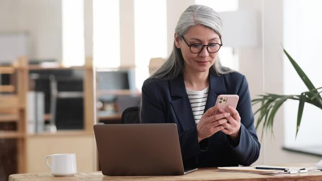 A confident asian woman in a glasses with gray hair typing something on the phone and then on the laptop in the office