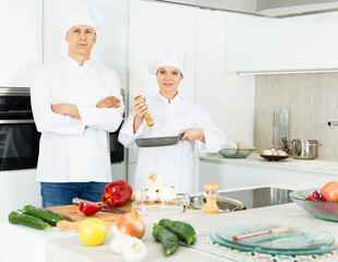 Two cheerful woman and man young chefs in uniform preparing food on kitchen