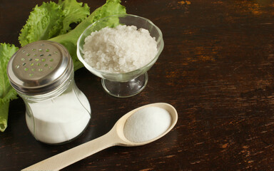 Glass salt shaker with salt, wooden spoon with salt and a vase with rock sea salt on a dark wooden background